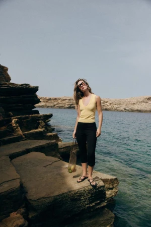 Model wearing yellow sleeveless top and black trousers standing on rocky coast holding string bag by ocean water