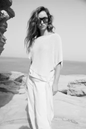 Model in oversized light knit top and sunglasses standing on a rocky coastal shore in a monochrome fashion photograph.