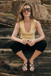 Model wearing dark sunglasses, yellow tank top, and black leggings squatting on layered rock formation.
