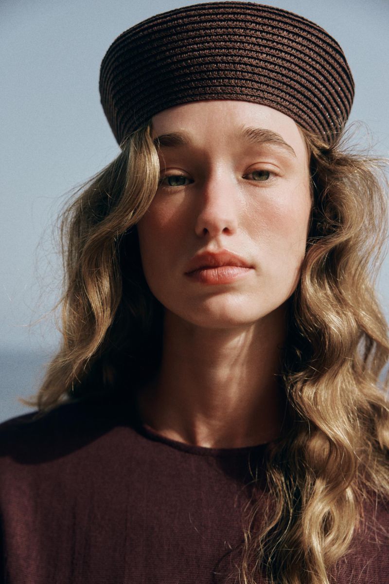 Close-up portrait of a model wearing a brown hat and dark top against a clear sky background