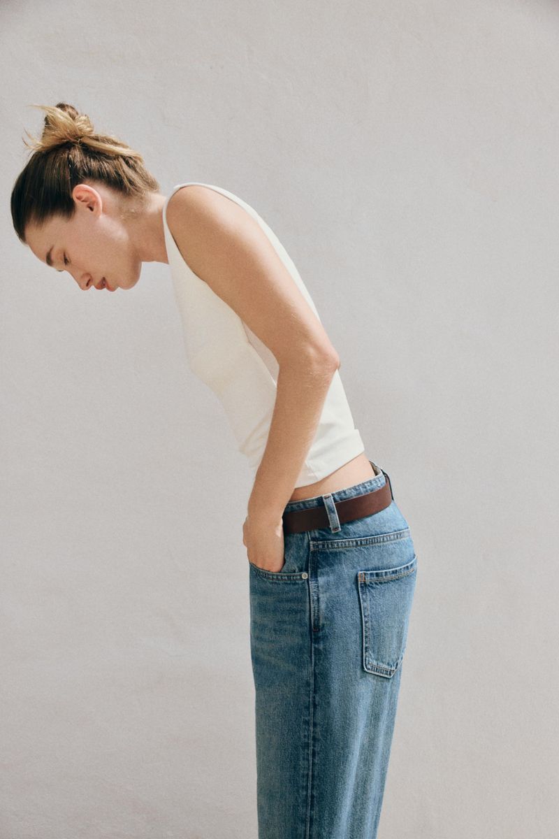 Profile view of a woman wearing a white top and blue jeans leaning forward against a light grey background