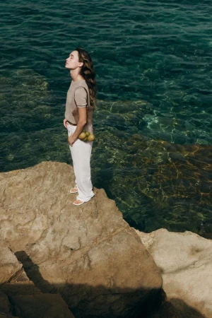 Model Jenn du Puy standing on coastal rocks overlooking the ocean holding a bag of lemons for Zara.