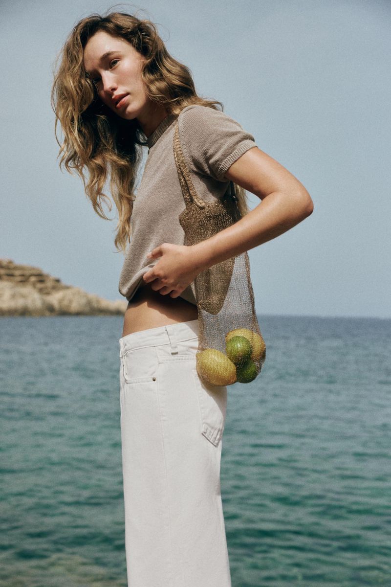 Model standing by the ocean wearing a brown knit top and white trousers holding a mesh bag with fruit