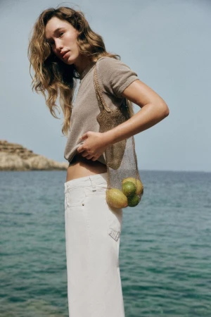 Model standing by the ocean wearing a brown knit top and white trousers holding a mesh bag with fruit