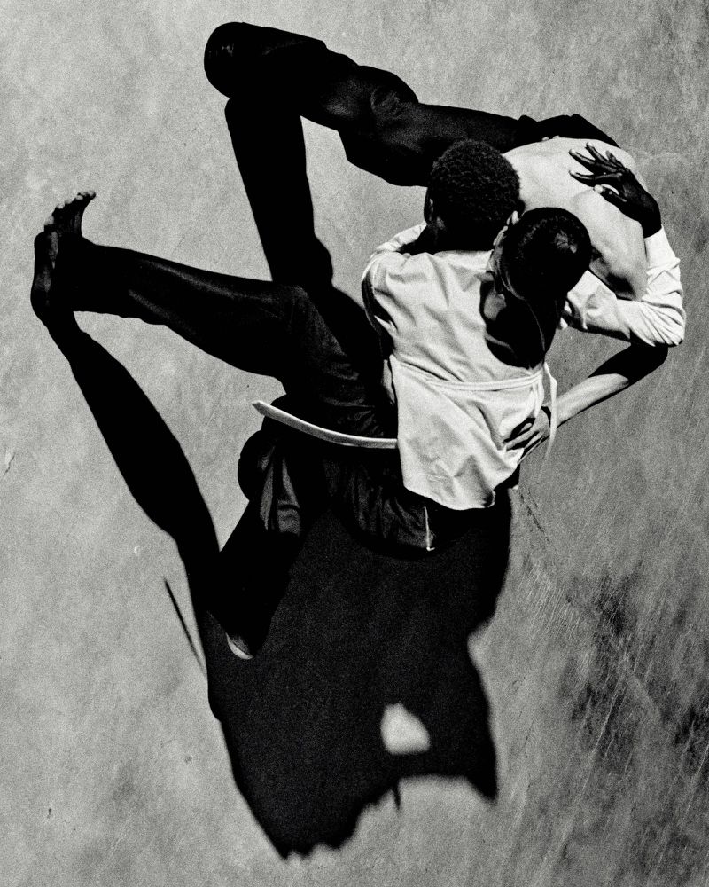 High-angle black and white shot of two men embracing in a choreographed dance or movement, casting long shadows on the ground.