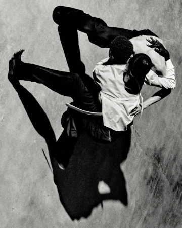High-angle black and white shot of two men embracing in a choreographed dance or movement, casting long shadows on the ground.