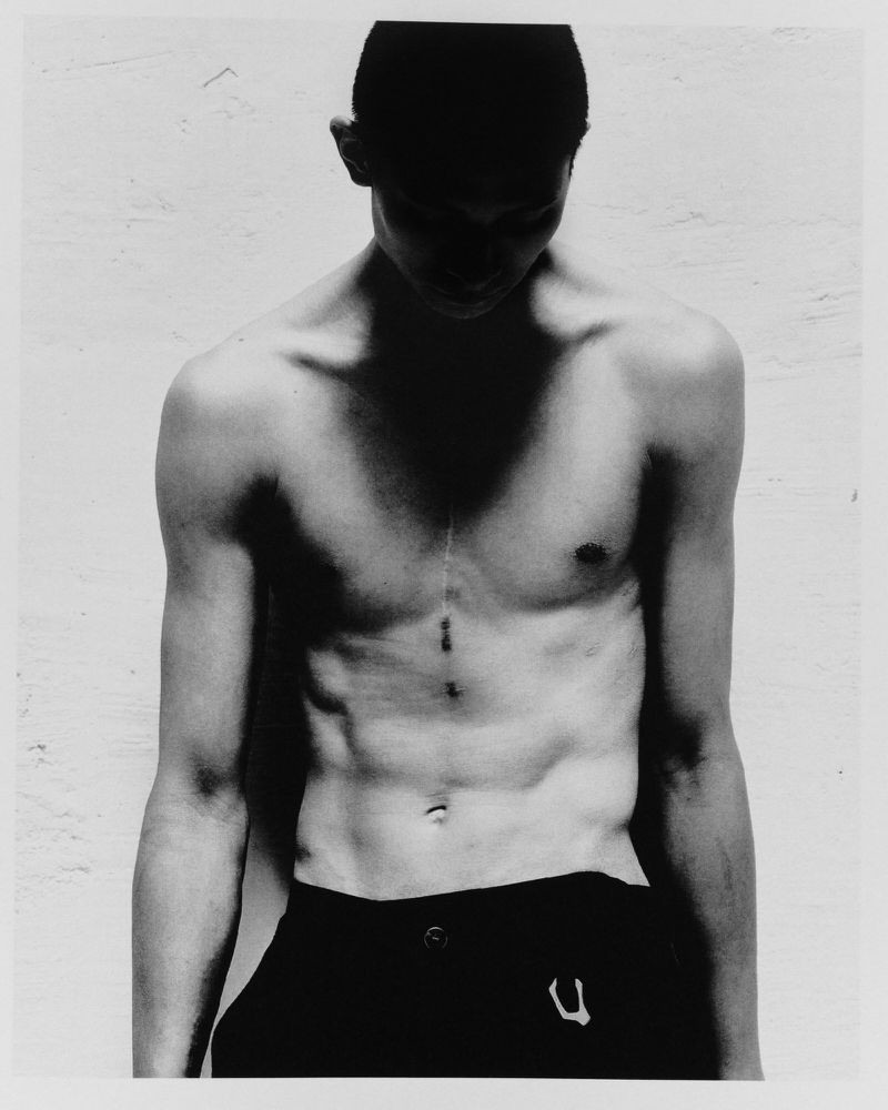 Black and white portrait of a shirtless adult male with dark hair and athletic build, looking down against a textured wall backdrop.