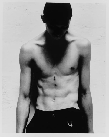 Black and white portrait of a shirtless adult male with dark hair and athletic build, looking down against a textured wall backdrop.