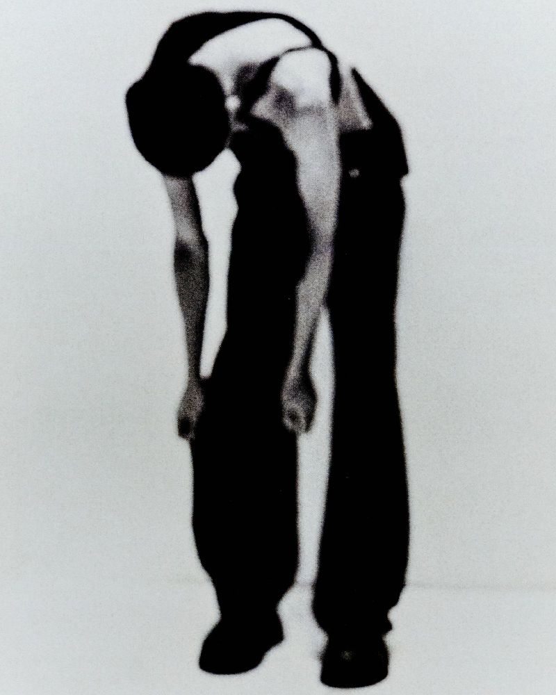 Monochrome full shot of an adult in black clothing bending over, facing the camera on a white background in a studio.