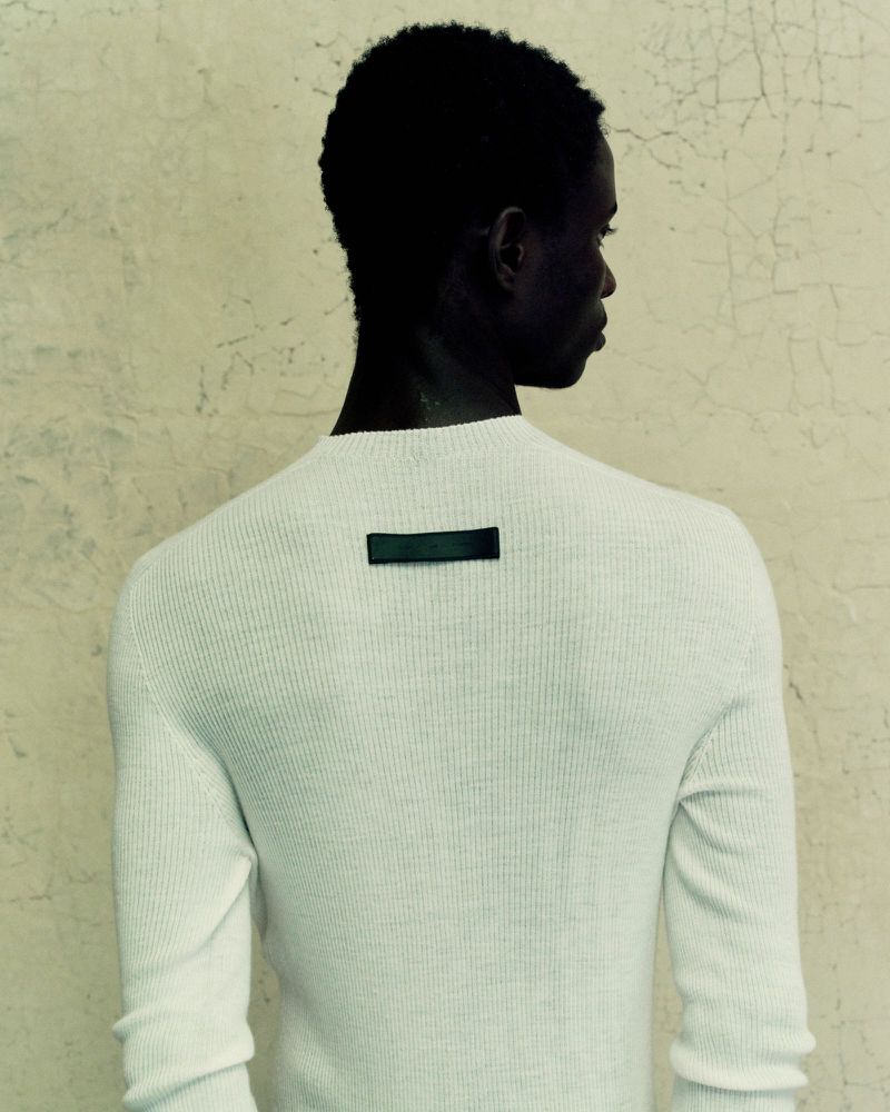 Back view of Black male in a white ribbed long sleeve sweater against a textured wall. The sweater has a dark rectangular label.