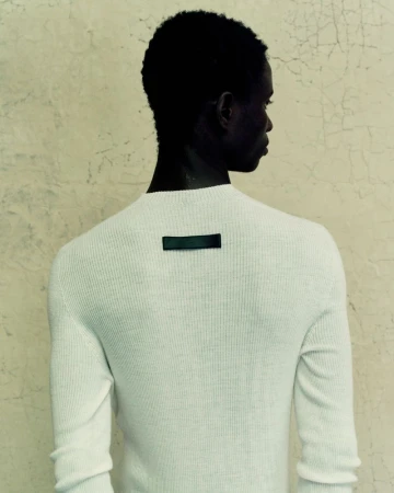 Back view of Black male in a white ribbed long sleeve sweater against a textured wall. The sweater has a dark rectangular label.