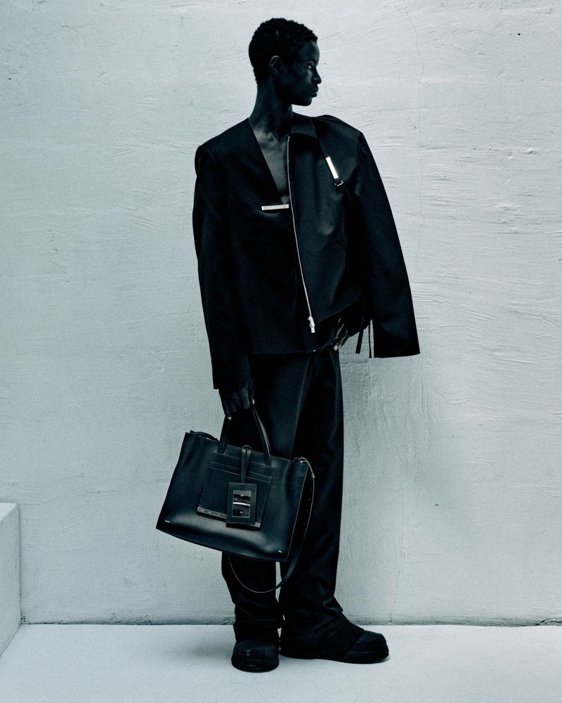 A slender Black model in a stylish black suit and chunky boots poses in a minimalist photo studio, holding a sleek black leather tote bag.