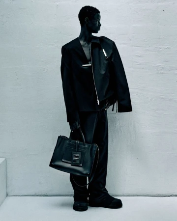 A slender Black model in a stylish black suit and chunky boots poses in a minimalist photo studio, holding a sleek black leather tote bag.