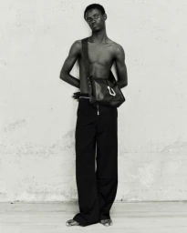 Shirtless Black male model wearing black wide-leg trousers and a leather crossbody bag against a textured wall.