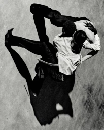 High-angle black and white shot of two models intertwined in a dynamic movement pose casting a sharp shadow on concrete.