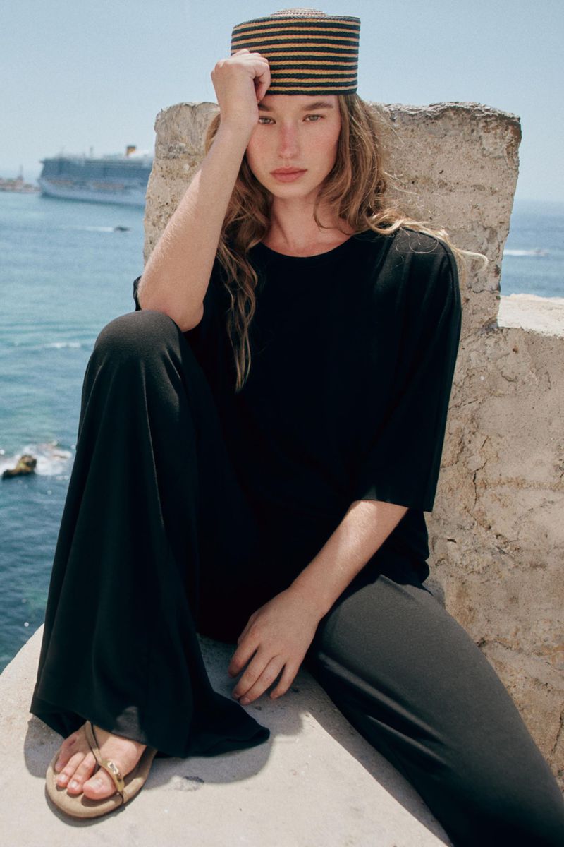 Model Jenn Du Puy seated on a stone wall by the sea wearing a black outfit and striped hat, photographed by Paolo Steve for Zara.