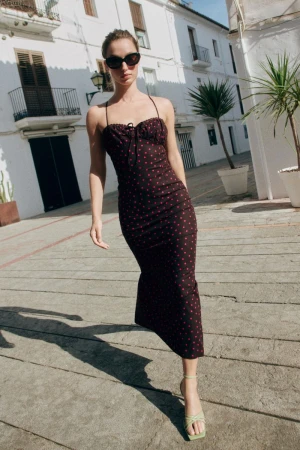 Model in dark sunglasses wearing a black polka dot dress walking on a cobblestone street by white buildings.