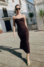Model in dark sunglasses wearing a black polka dot dress walking on a cobblestone street by white buildings.