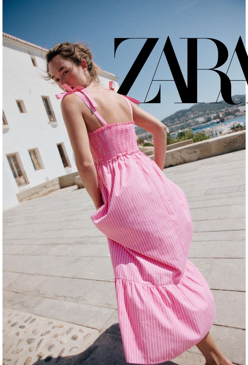Model wearing a pink and white striped midi dress walking on a stone paved terrace against a white building under a clear sky.