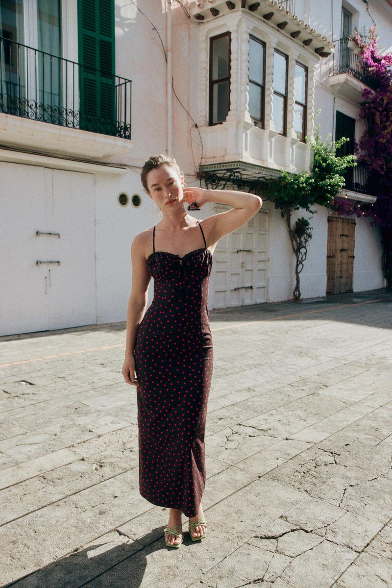 Model Jenn du Puy wearing a dark polka-dot slip dress posing on a sunlit European cobblestone street with white buildings.