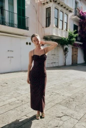 Model Jenn du Puy wearing a dark polka-dot slip dress posing on a sunlit European cobblestone street with white buildings.