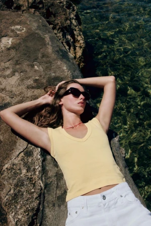 Model wearing dark sunglasses and a yellow tank top lying on rocky coast by clear water
