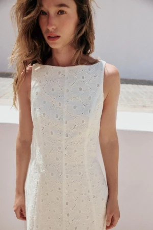 Model wearing a white eyelet broderie anglaise sleeveless dress outdoors against a bright sunlit architectural wall
