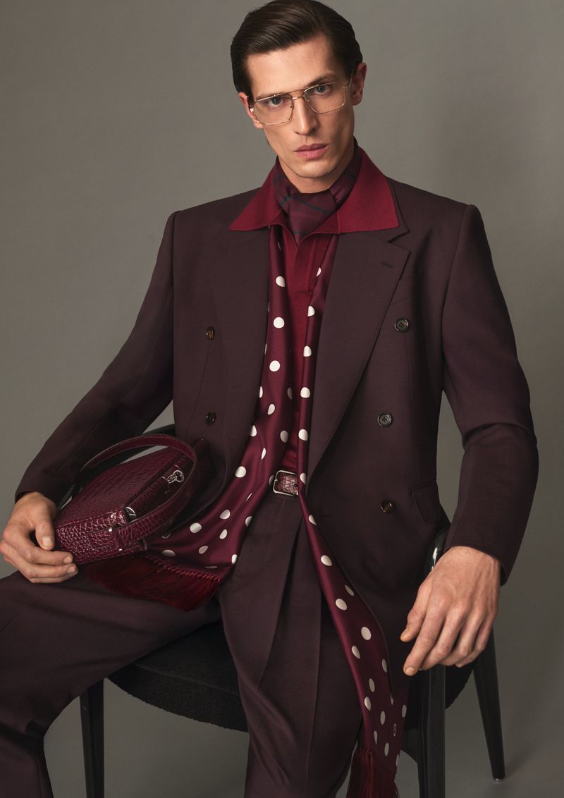 Seated male model wearing a maroon double-breasted suit holding a structured handbag against a grey backdrop