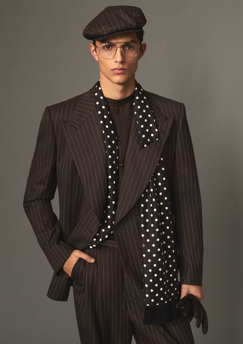 Male model wearing a pinstripe suit, flat cap, glasses, and polka dot scarf against a solid grey background