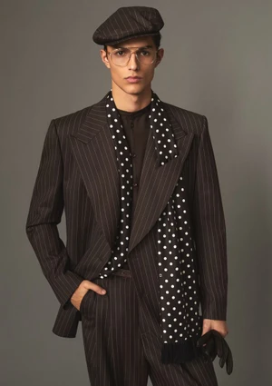 Male model wearing a pinstripe suit, flat cap, glasses, and polka dot scarf against a solid grey background