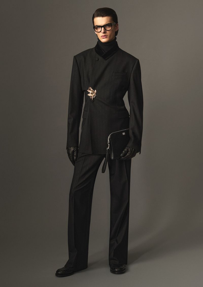 Model in black suit holding a clutch and wearing glasses against a grey seamless studio backdrop.