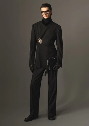 Model in black suit holding a clutch and wearing glasses against a grey seamless studio backdrop.
