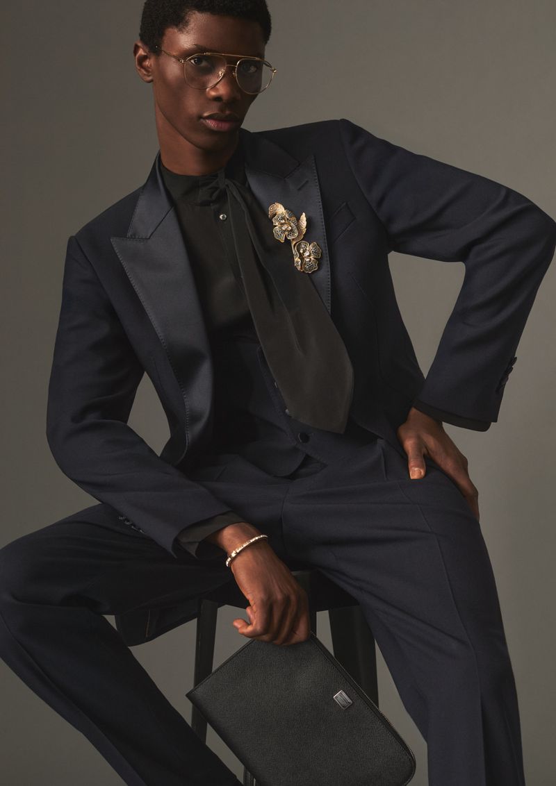 Model seated wearing a dark navy suit, black shirt, tie, and glasses while holding a dark clutch against a grey studio backdrop.