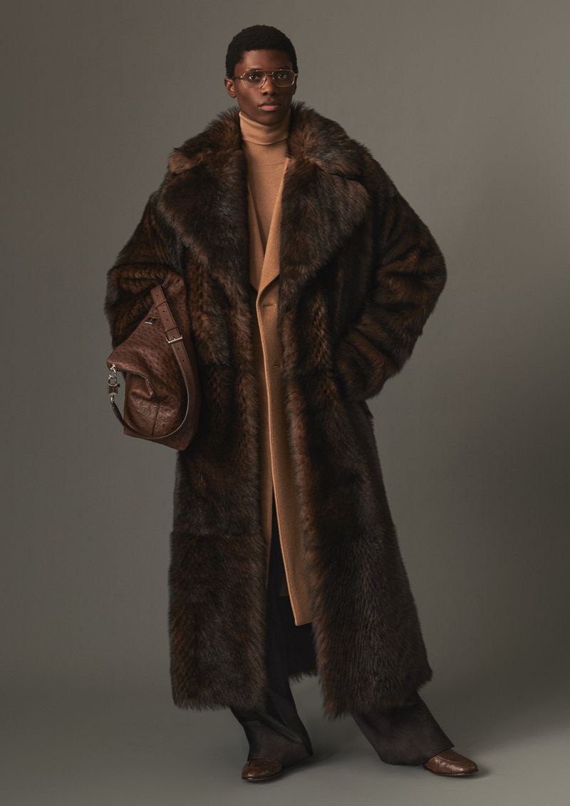 Model wearing a long brown fur coat and carrying a leather handbag in a studio