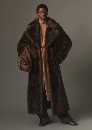 Model wearing a long brown fur coat and carrying a leather handbag in a studio