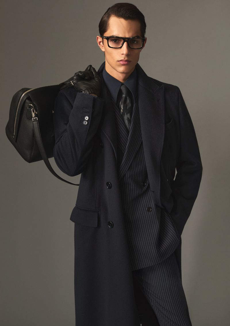 Man wearing dark coat and glasses holding a black leather travel bag against a neutral studio background