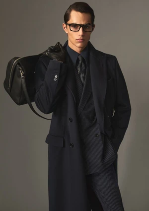 Man wearing dark coat and glasses holding a black leather travel bag against a neutral studio background