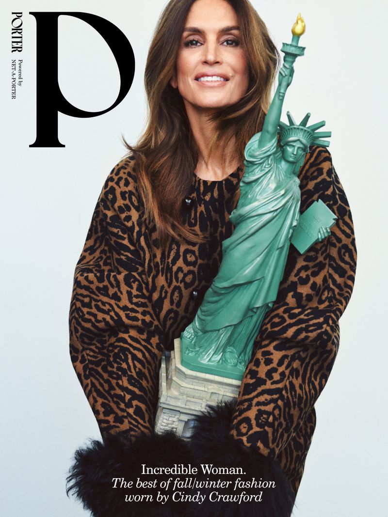 Cindy Crawford holding a miniature Statue of Liberty wearing a leopard print coat against a white background