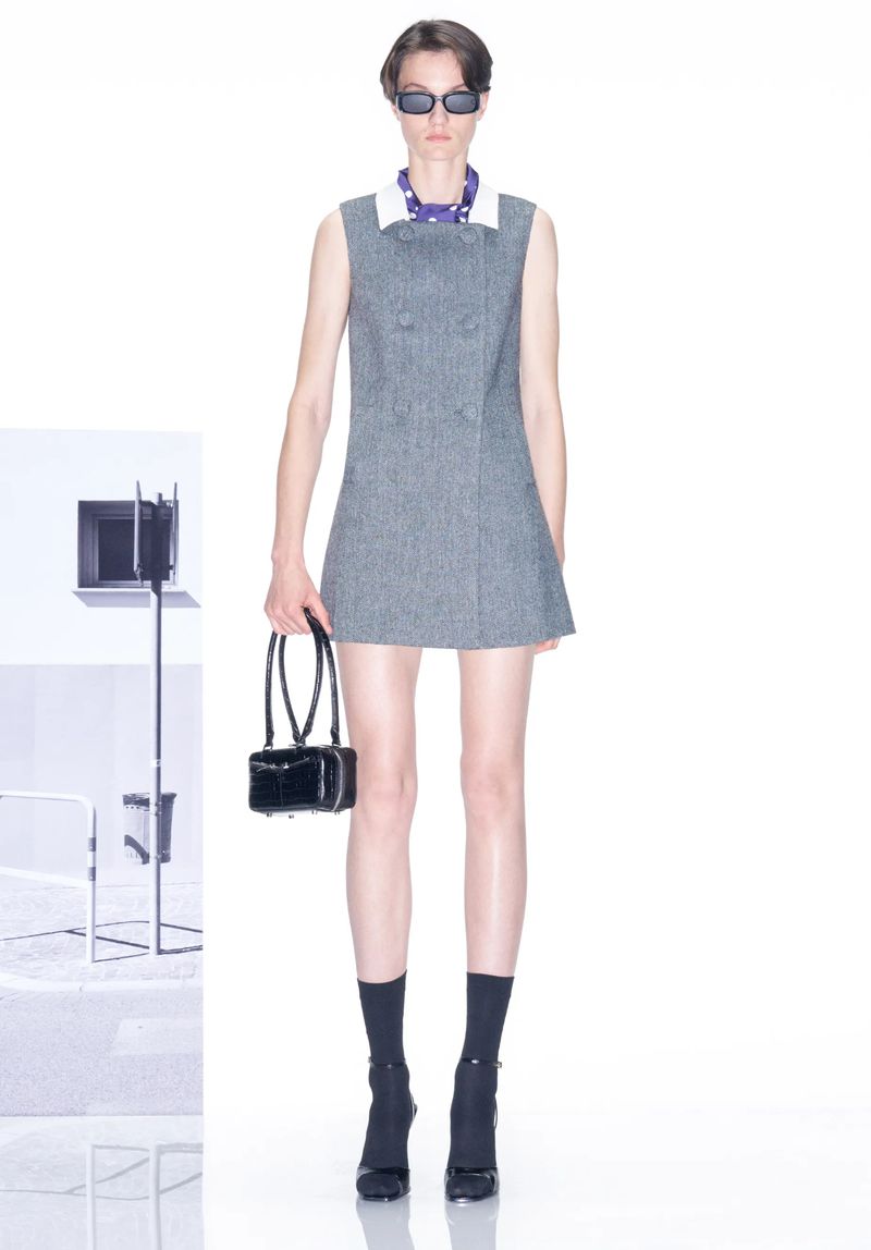 Fashion model wears grey mini dress with black accessories in bright studio. Studio lighting setup visible in soft focus background.