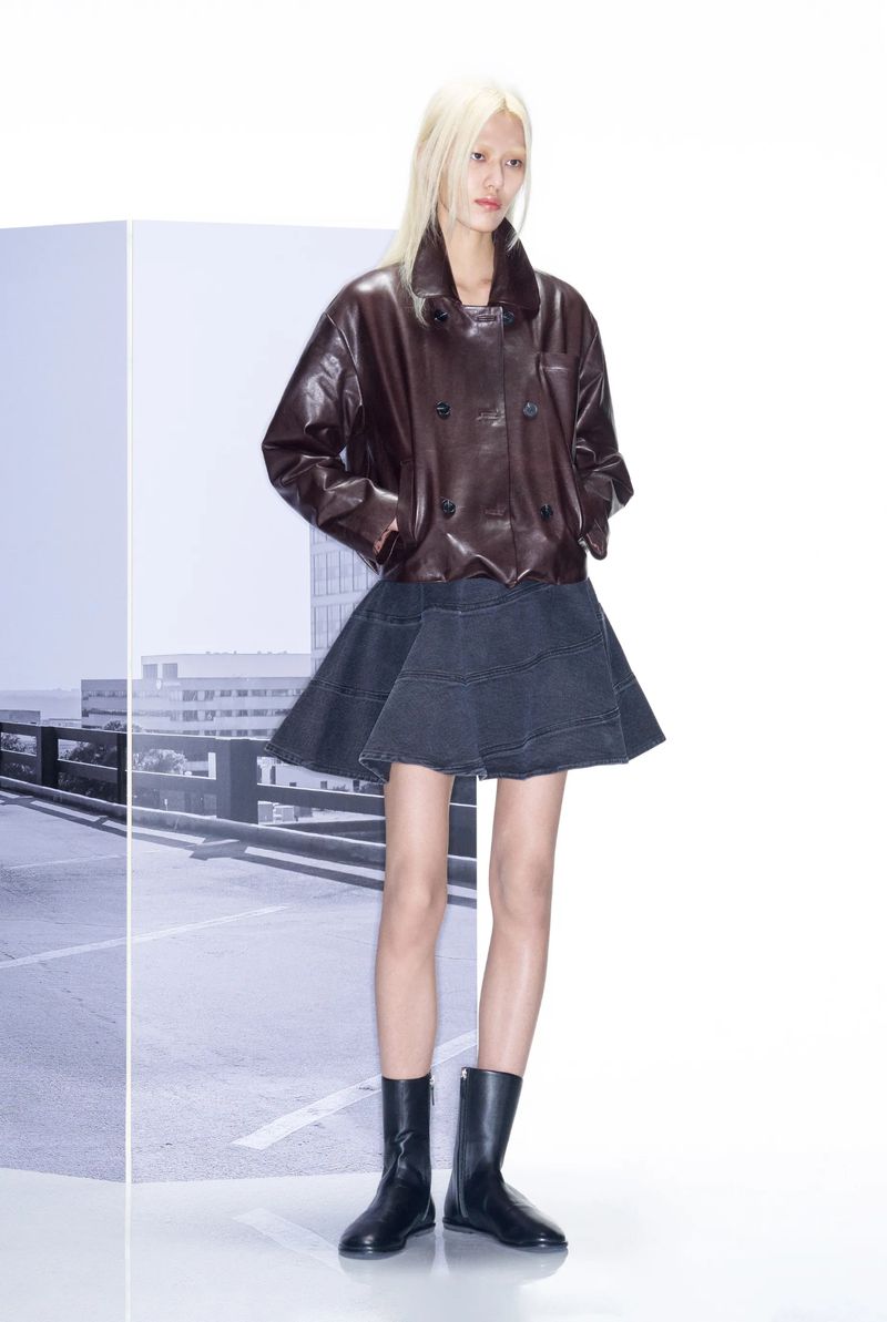 A slender fashion model poses in a brown leather jacket, dark skirt, and black boots against a bright studio backdrop.