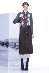 Model wearing argyle sweater and brown skirt holding handbag against a white studio background