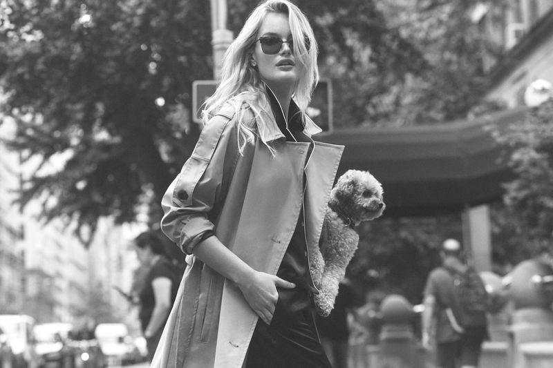 Model wearing dark sunglasses and a belted coat outdoors while holding a small dog in a street setting.