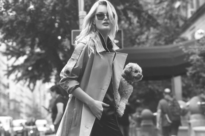 Model wearing dark sunglasses and a belted coat outdoors while holding a small dog in a street setting.