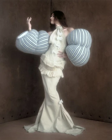Model wearing a structured cream gown with voluminous striped puffed sleeves against a mottled dark brown corner backdrop