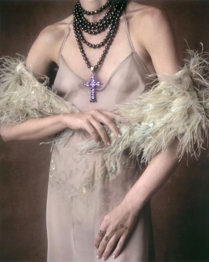 Model wearing a neutral slip dress, feathered stole, pearl necklace with large cross pendant, and tattoos on fingers.