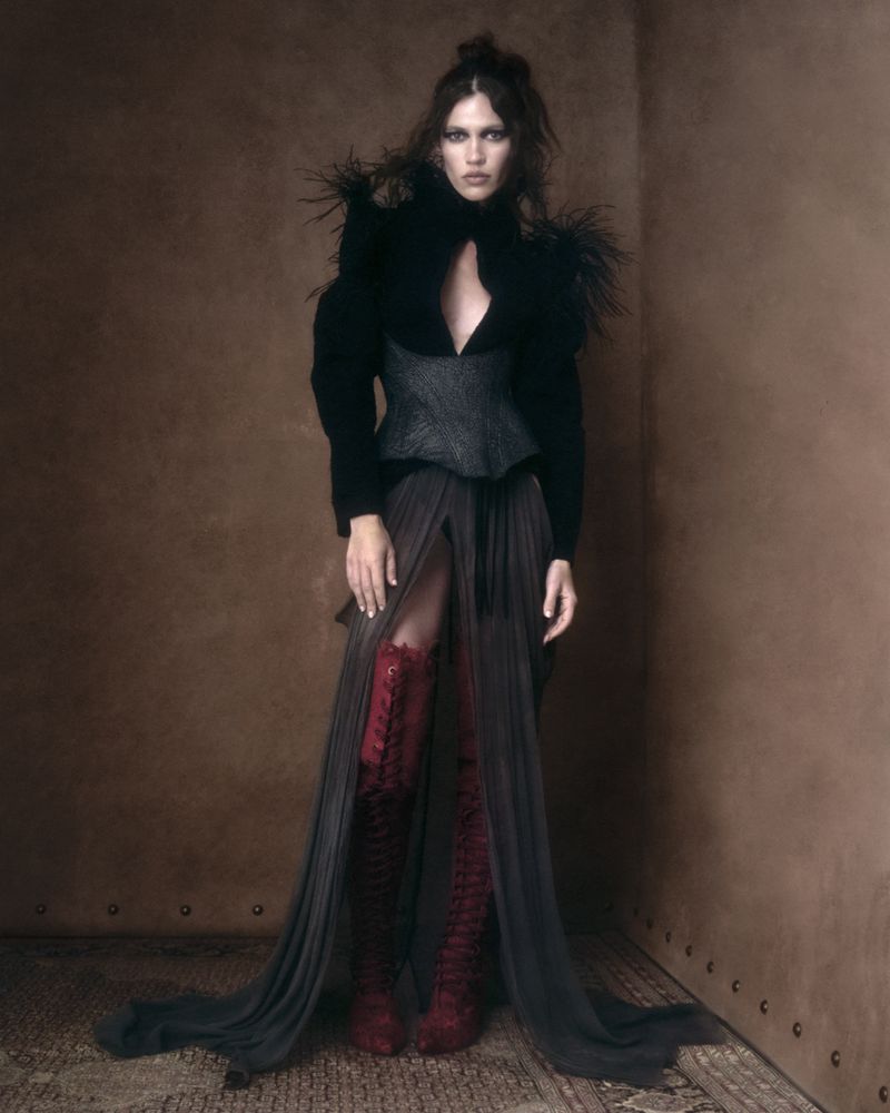 Model in black feathered top and long sheer skirt standing against a brown textured wall in an editorial photograph