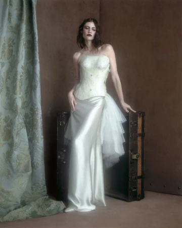 Model in strapless white gown and corset posing against a brown wall next to a vintage trunk and draped curtain