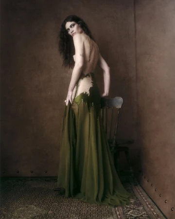 Model wearing a backless olive green gown, standing beside a wooden chair against a dark textured studio backdrop.