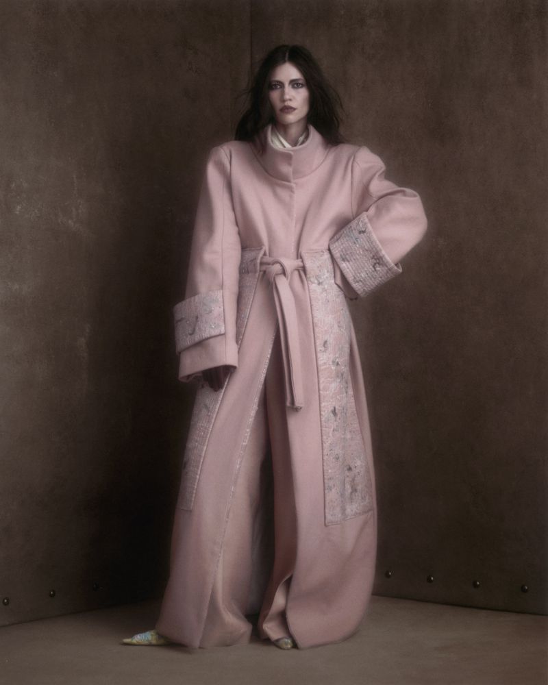 Model standing against a textured brown wall wearing a long pink coat with patterned panels and matching trousers.