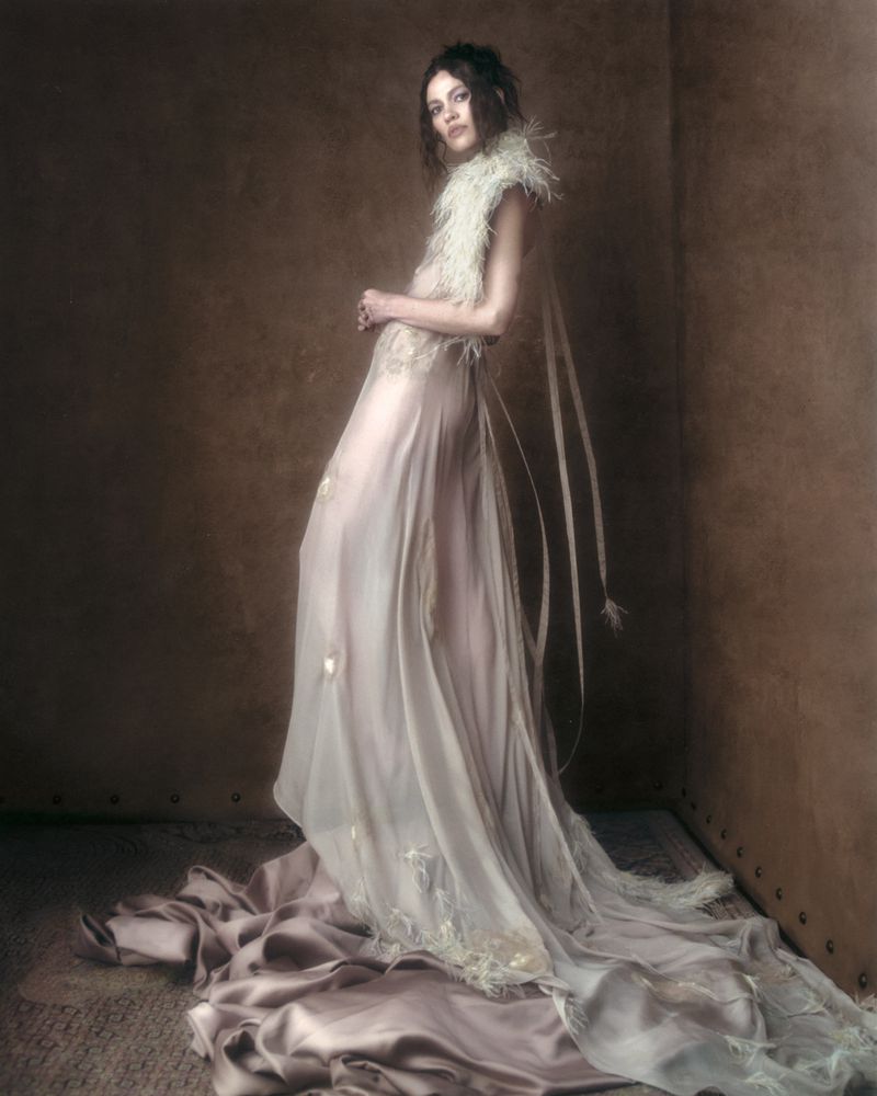 Model in a flowing white gown with feathered neckline posing against a dark textured studio wall.
